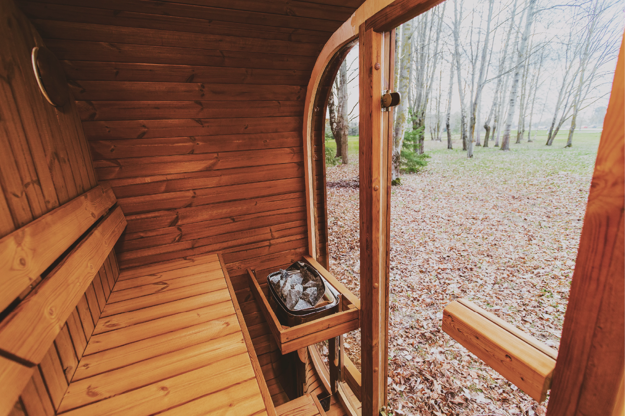 Interior of a Mande Spa USA MW16 outdoor sauna made from sustainably sourced thermowood, showing wooden benches and a sauna heater with stones, with a view of a leafy forest through the open door