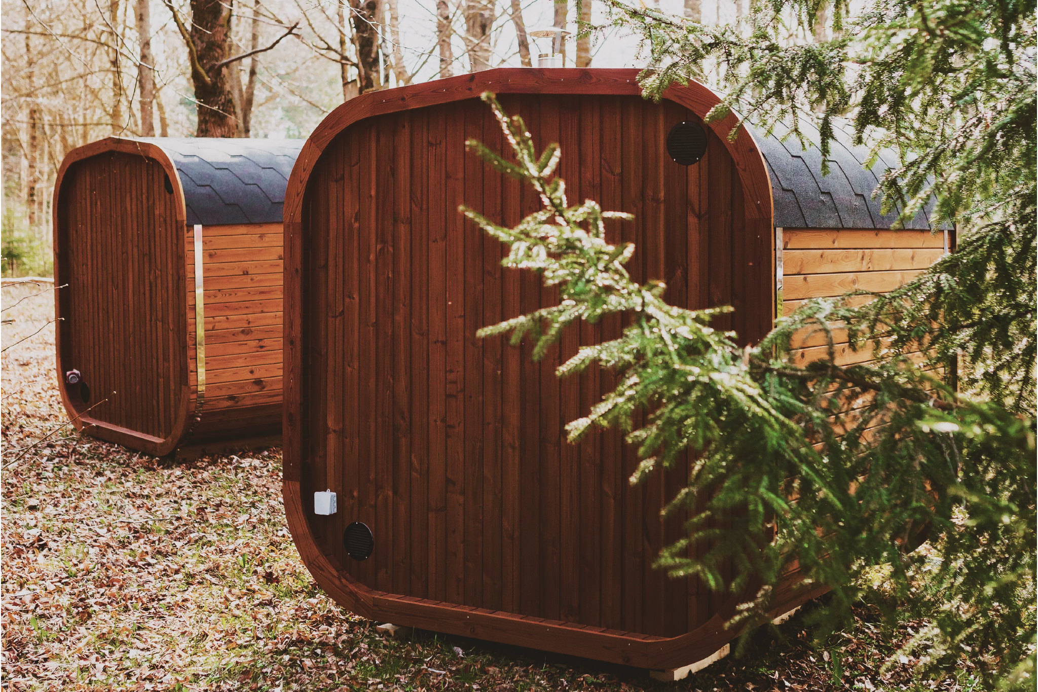 Back view of two Mande Spa USA MW16 outdoor saunas made from sustainably sourced thermowood, set on a forest floor with fallen leaves and surrounded by trees